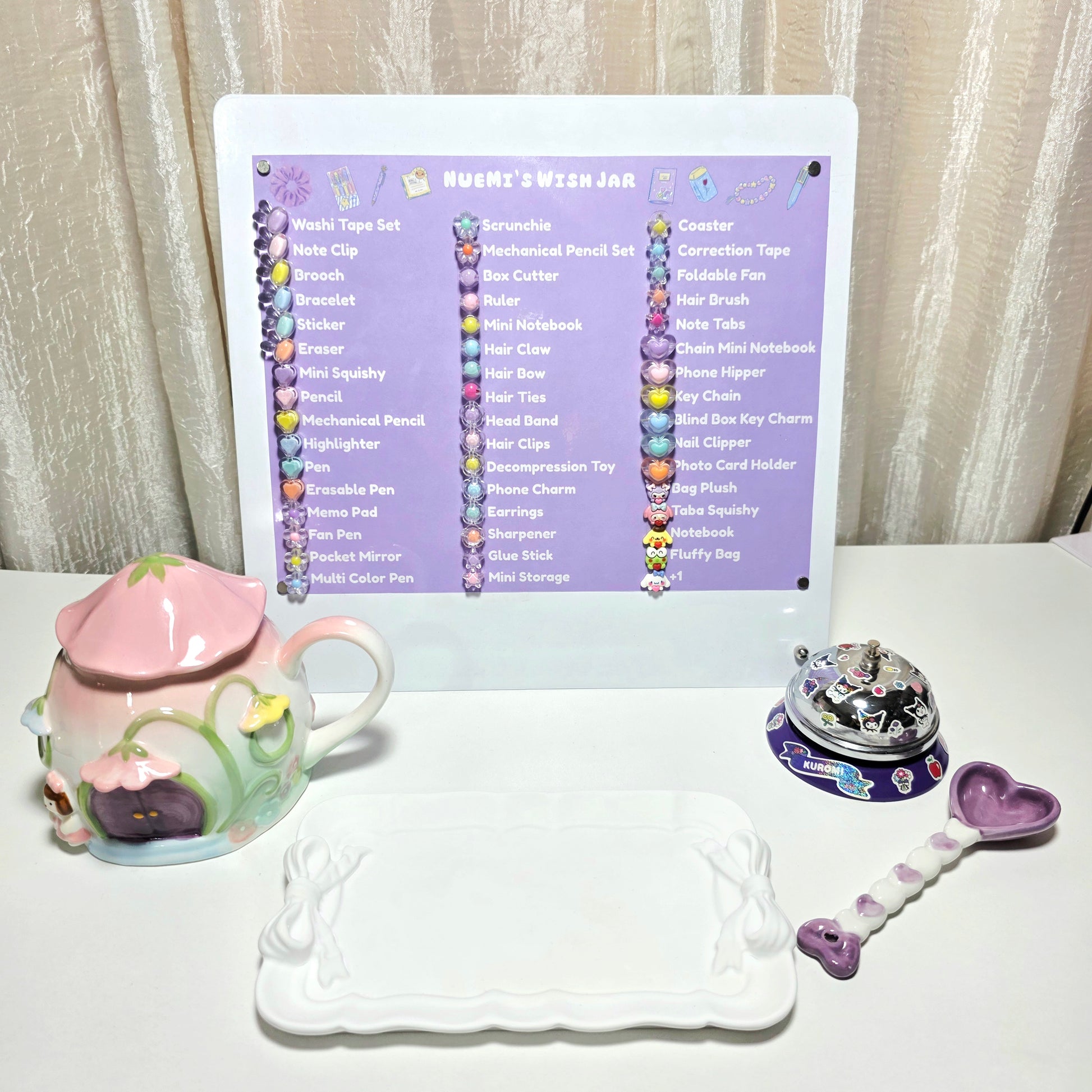 A tabletop display of a white desk calendar titled 'NUEMI'S WISH JAR' with a purple header and colorful decorative elements, surrounded by themed accessories including a pink ceramic mug, a white tray, a small round desk bell, and a purple heart-shaped ceramic spoon, set against a beige curtain background.