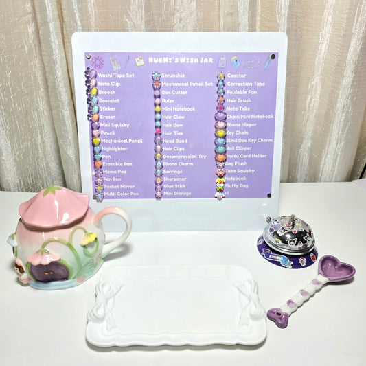 A tabletop display of a white desk calendar titled 'NUEMI'S WISH JAR' with a purple header and colorful decorative elements, surrounded by themed accessories including a pink ceramic mug, a white tray, a small round desk bell, and a purple heart-shaped ceramic spoon, set against a beige curtain background.